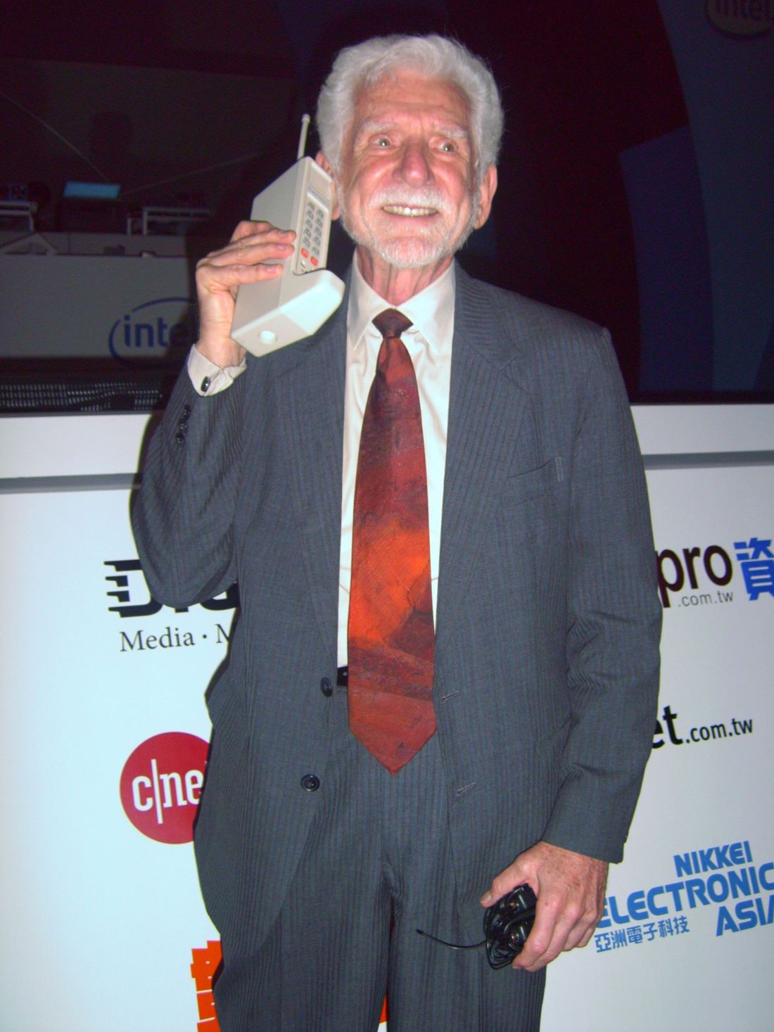 Martin Cooper in a suit and red tie holding a large DynaTAC prototype phone to his ear at a tech forum in 2007