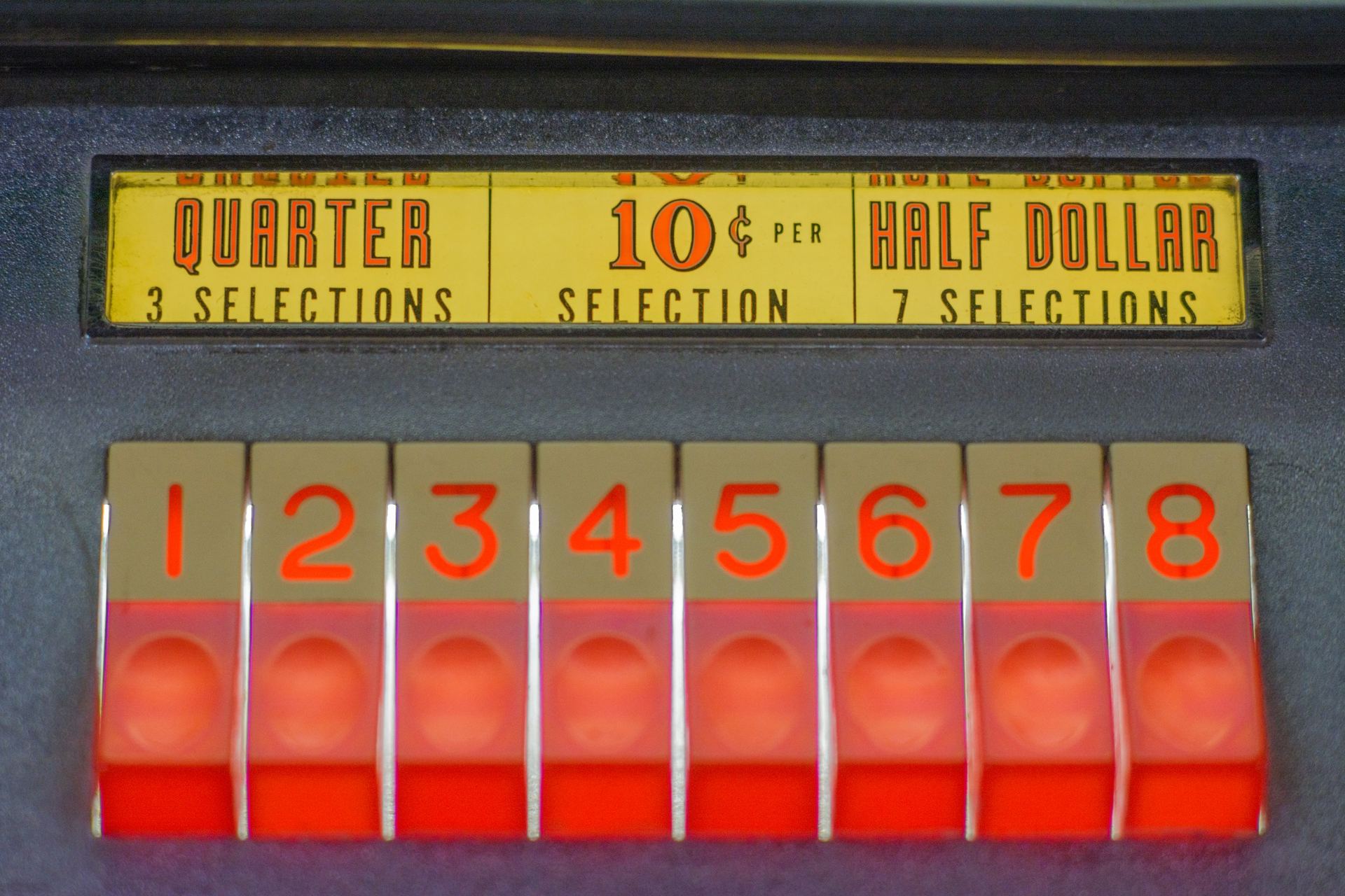Close-up of glowing red numbered selection buttons on a vintage Seeburg jukebox, under a yellow price panel reading quarter, 10 cents per selection and half dollar