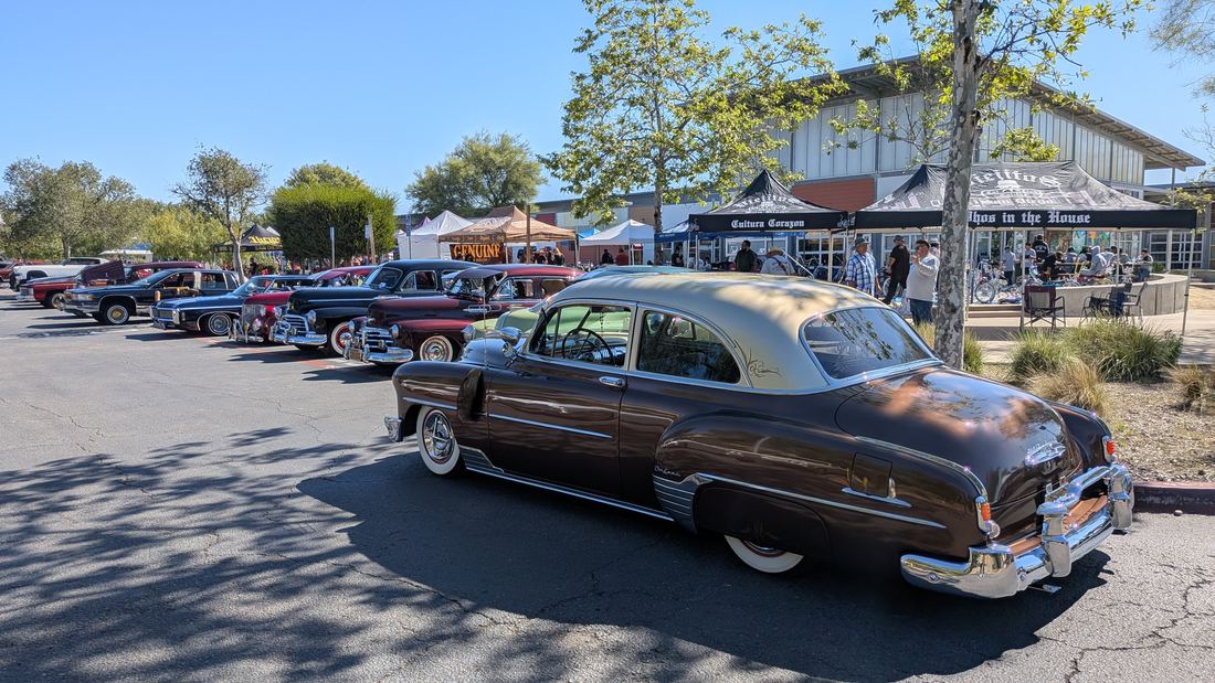 A two-tone brown and cream vintage lowrider sedan with whitewall tires parked in front of a row of classic cars at a car show in Chula Vista, California