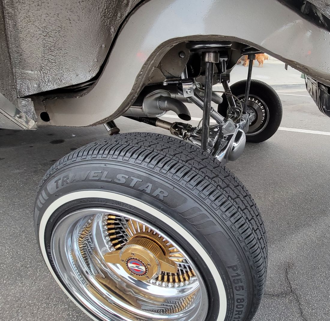 Close-up of a lowrider's raised front wheel showing a whitewall tire, a gold and chrome wire wheel and the suspension arms of the lifting system
