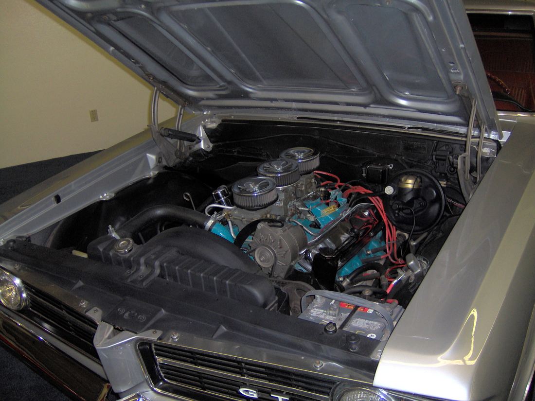 The open hood of a silver 1964 Pontiac GTO showing a blue 389 V8 engine topped with three round chrome air cleaners