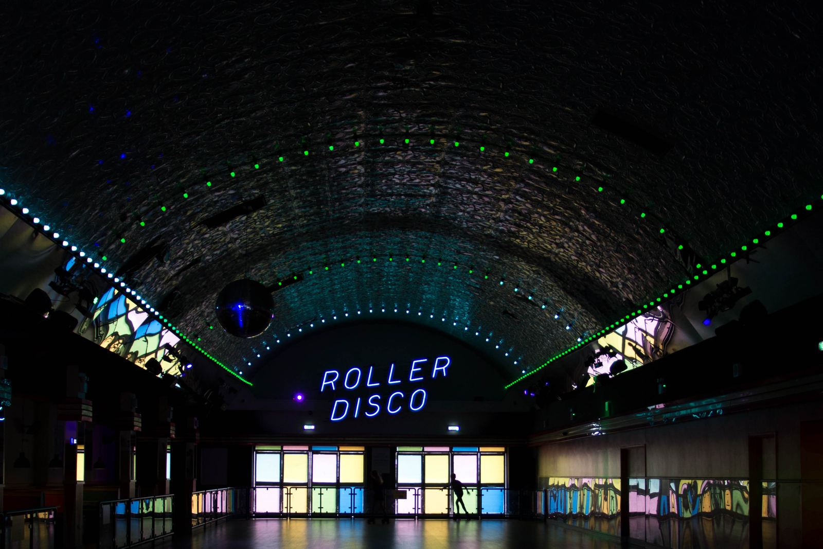 A dark arched roller rink hall lit with green laser lights, a mirror ball and a glowing neon Roller Disco sign