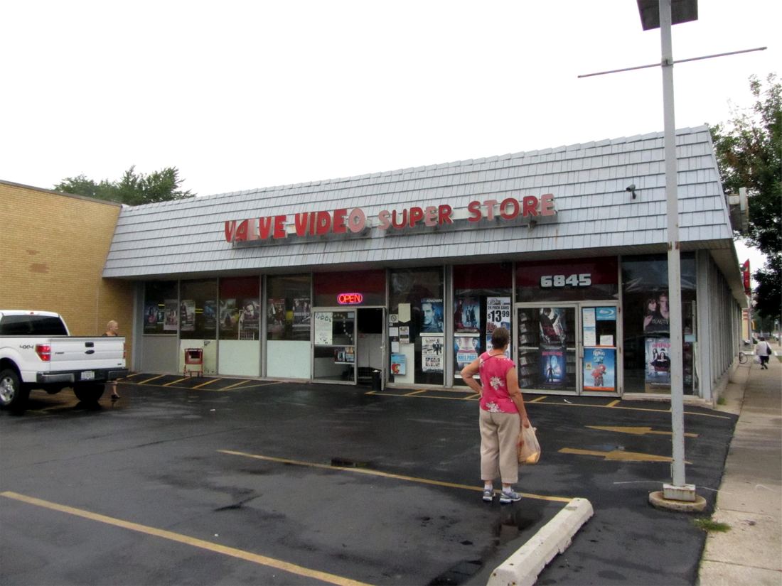 A neighborhood video rental store in a small strip building with a red sign and a customer walking toward the door